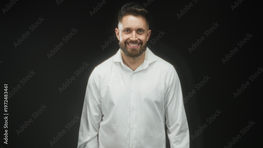 Obraz premium Man with beard smiling, hands at sides in studio wearing white shirt and collar visible; friendly approachable presence.