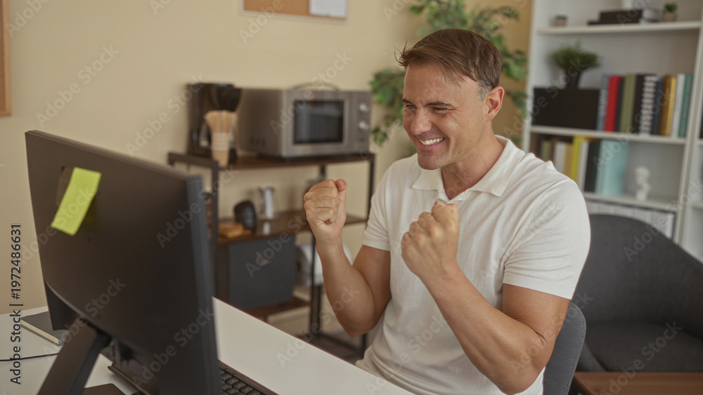 Fototapeta premium Man clenches fists and smiles at computer screen with sticky note on monitor in building office, leaning forward at desk; excitement triumph.