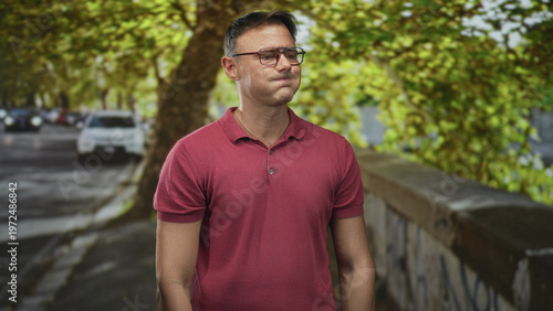 Man in red polo wearing glasses looking up on a street by a graffiti wall near parked cars and leafy trees; contemplation solitude.