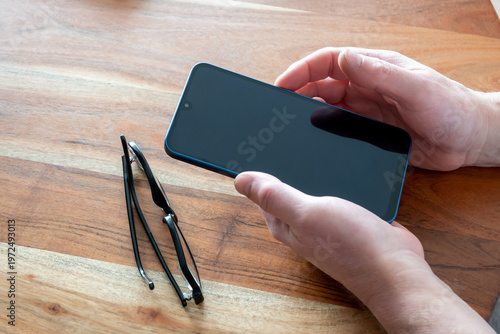 Hand holding new mobile phone on the wood table. Work in office with smartphone and glasses.