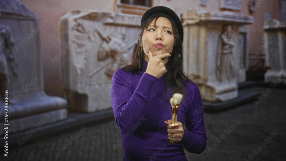 Obraz premium Woman holding makeup brushes, finger to chin gesture, posed before stone sculptures in building; quiet contemplation.