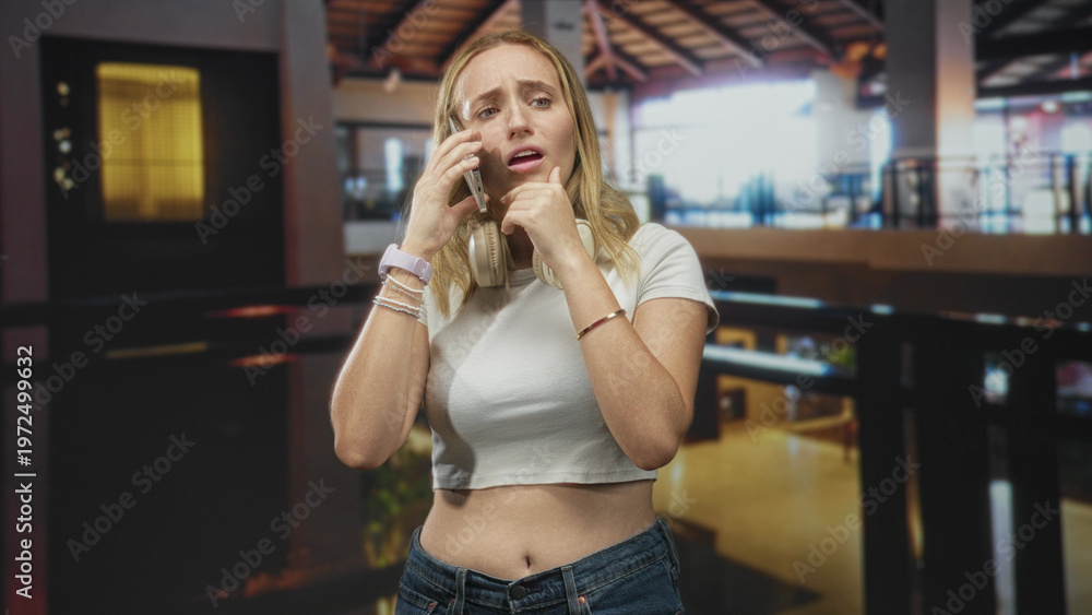 Fototapeta premium Woman wearing headphones holds phone to ear with hand on chin and exposed stomach in an airport terminal; anxiety concern.