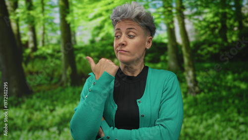 Obraz na plátne Woman with short gray hair wearing green cardigan and black top points thumb at forest backdrop indoors; skepticism doubt cynicism distrust