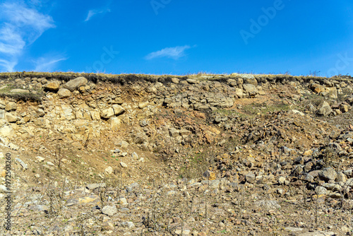 A cross-section of a hill with visible geology. Layers of large rocks, small gravel, and brown earth. A layer of yellow grass on the summit. A bright blue sky in the background.