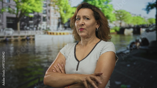 Woman with arms crossed on street by amsterdam canal building facade under sunlight; confidence and resilience.