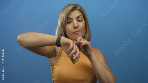 Fotografie Woman wearing orange sleeveless top pointing finger to watch on bare wrist in blue studio; impatience time pressure urgency
