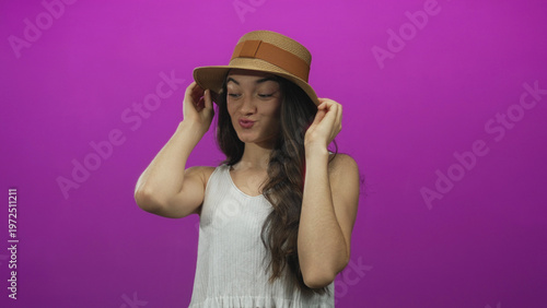 Foto Young hispanic woman adjusting straw hat with closed eyes in purple studio; playful excitement
