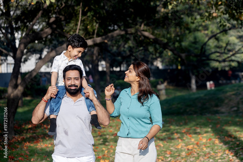 Indian parents walking with their child sitting on father shoulders in a park. Joyful family bonding and lifestyle moment.