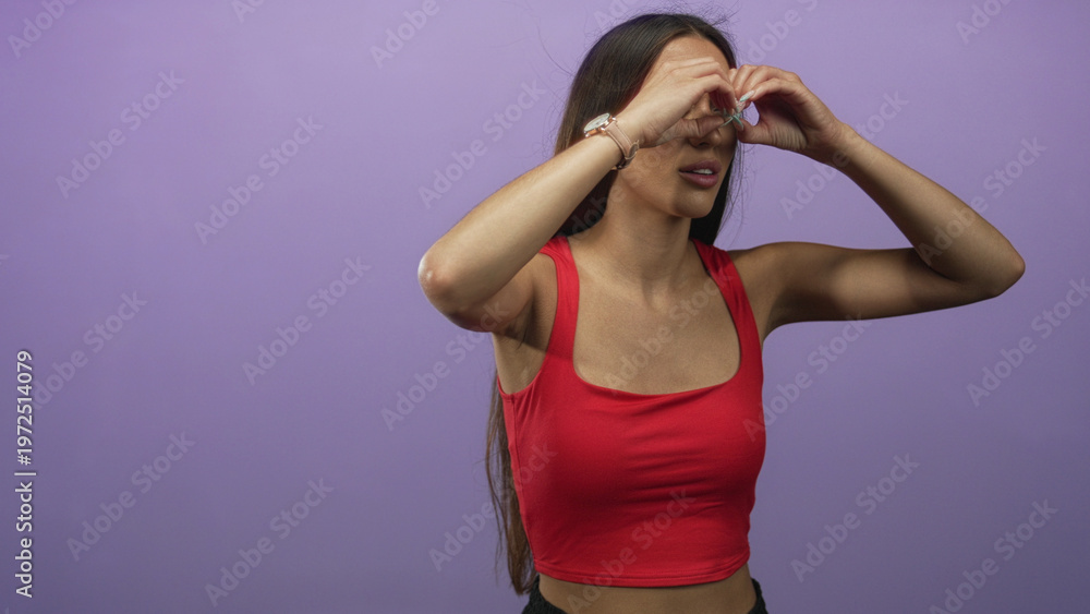 Fototapeta premium Woman wearing red crop top, bare arms visible, forms heart with hands over eyes in studio with purple backdrop; playful affection.