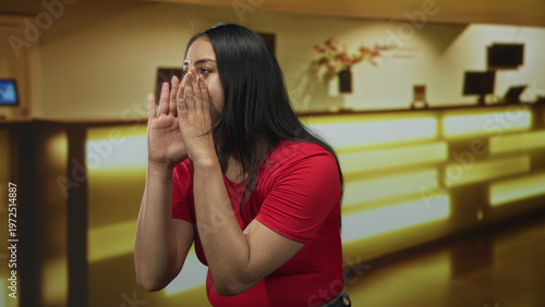Принти на полотні Young hispanic woman cups hands to mouth calling for help at hotel front desk building; concern urgency