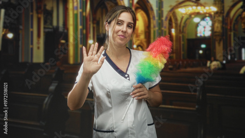 фотографія Woman in cleaning uniform holding rainbow feather duster waves right hand inside church building with pews and stained glass; welcoming warmth