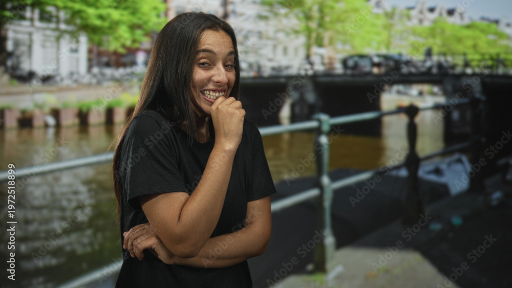 Fototapeta premium Woman laughing with fist to mouth and crossed arms standing by canal railing on an amsterdam street with bridge behind her; joy spontaneity.
