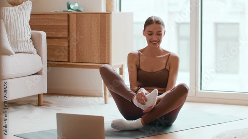 A woman with light skin is sitting cross-legged on a yoga mat at home. She is focused on her laptop and appears to be participating in an online fitness class, enjoying her workout.