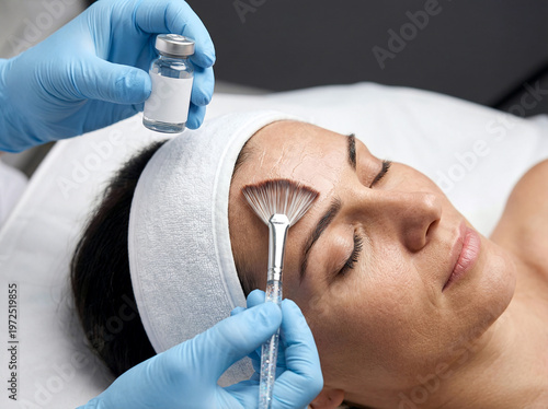 Esthetician in blue gloves applying a liquid facial treatment to a woman's forehead using a fan brush and a vial