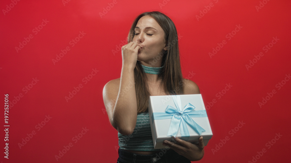 Fototapeta premium Young brunette woman holding white gift box with blue ribbon, smiling and waving her arm while presenting the present in studio; joyful celebration.