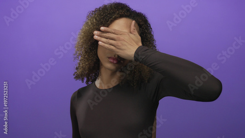 Obraz na plátně Woman covering her eyes with one hand in a purple studio and showing curly hair and pink lips; self reflection