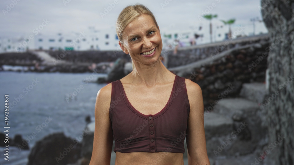Fototapeta premium Woman smiles and tilts head, showing exposed midriff and maroon tank top in front of a seaside building with volcanic rocks; joy.