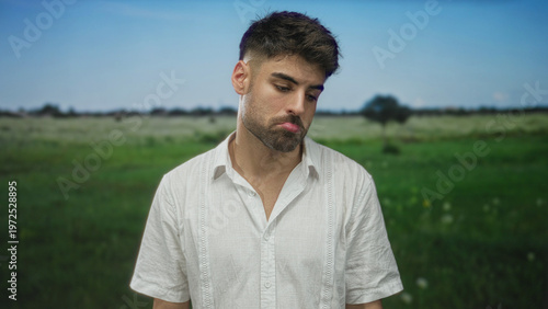 Man pursing lips in forest clearing under blue sky and green meadow in white linen shirt with soft sunlight filter; serenity.