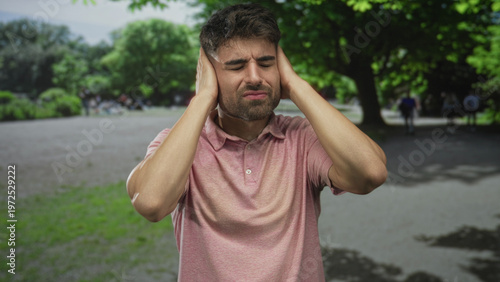 Quadro su tela Young hispanic man covering ears with both hands in forest framed by green trees and grass; annoyance