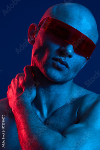 Stylish man with red sunglasses posing confidently against a vibrant blue background, showcasing a blend of modern fashion and artistic lighting.