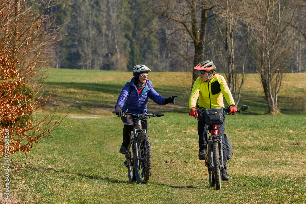 Obraz premium Happy senior couple enjoying an active lifestyle, riding electric bikes on a forest trail in spring. Outdoor recreation, healthy aging, and leisure time in nature on a sunny day.