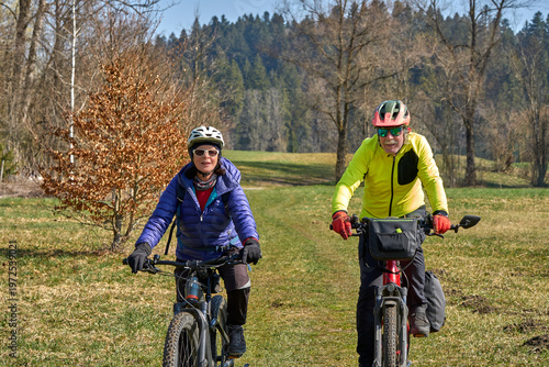 Happy senior couple enjoying an active lifestyle, riding electric bikes on a forest trail in spring. Outdoor recreation, healthy aging, and leisure time in nature on a sunny day.
