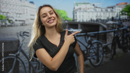Foto Young blonde woman finger pointing to the right on amsterdam street by canal and parked bicycles, smiling and looking sideways; playful confidence