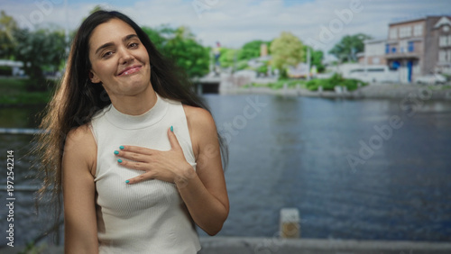Canvas-taulu Woman smiling with hand on chest wearing white top beside amsterdam canal water on city street; gratitude