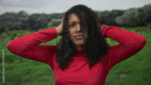 Photos Woman with curly hair in a red top covers her ears in a forest clearing; frustration discomfort silence
