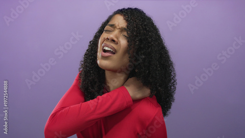 Obraz na plátne Curly haired woman in red top holds shoulder with a grimace in studio against purple background; pain