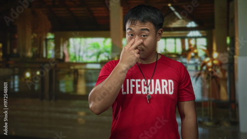 Fotografie Young man lifeguard wearing red shirt and holding whistle pinches nose with serious expression in hotel building; disgust