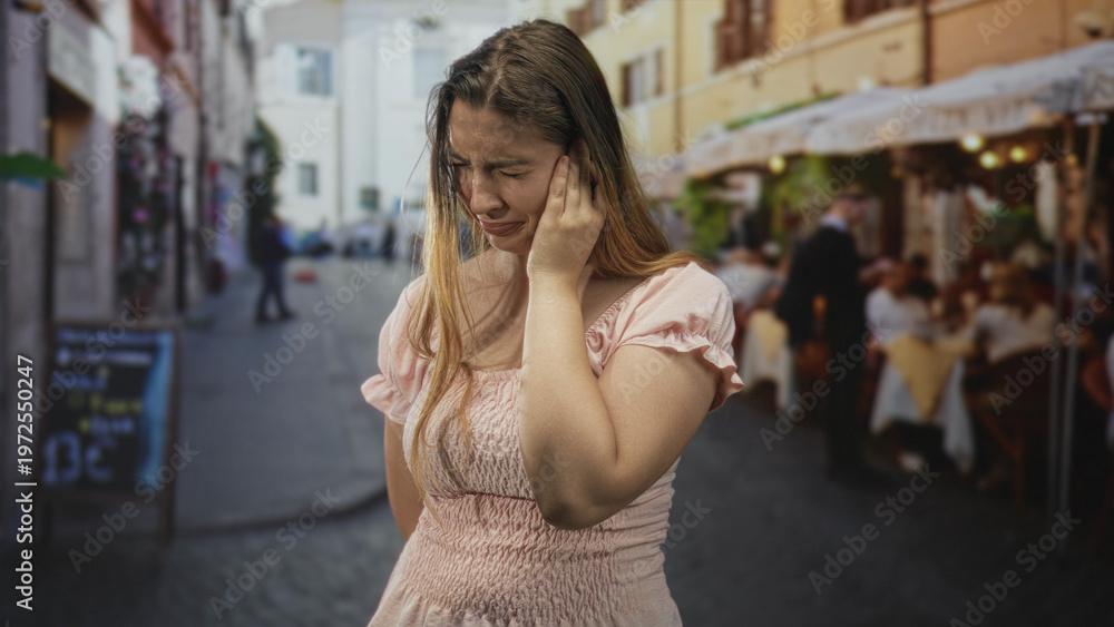 Fototapeta premium Woman holding her ear with one hand and wincing in visible pain at a busy street cafe with tables and diners; earache distress.