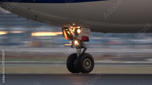 A close up, panning shot of an airplane nose landing gear with lights on. The wheels spin rapidly as the jet moves at high speed along the airport runway during takeoff