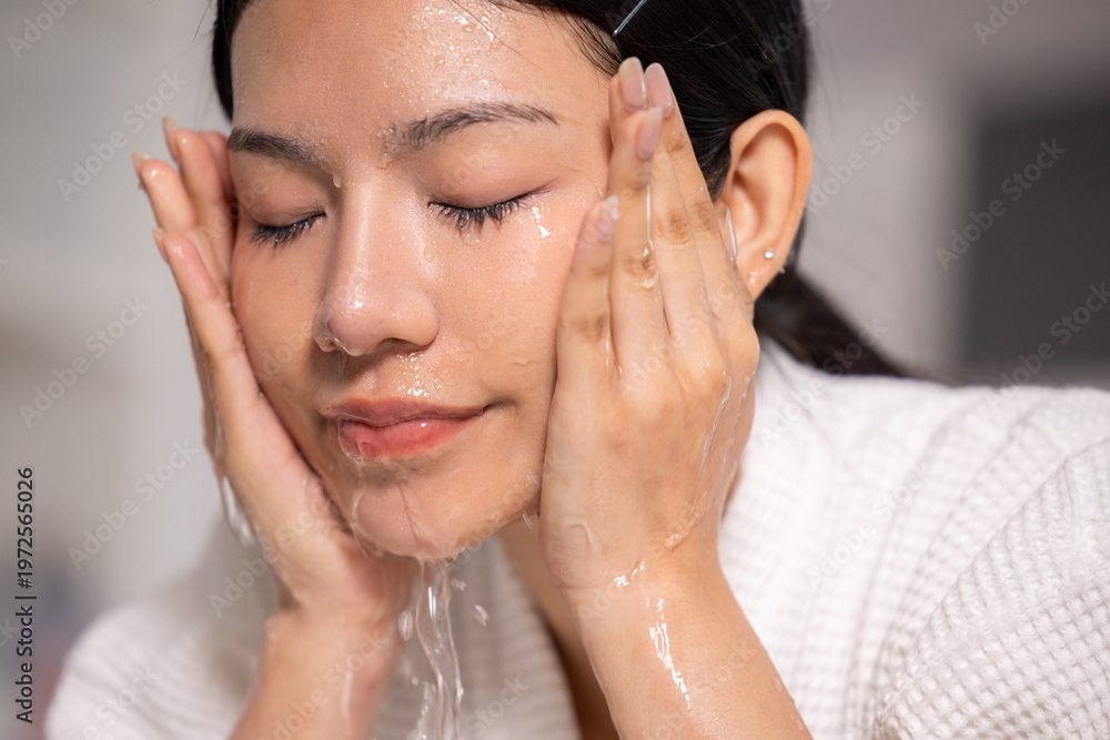 Naklejka premium Young Asian woman happily washing face with clear water during daily self care morning routine showing peaceful emotion enjoying refreshing skin cleaning treatment at home wearing white robe