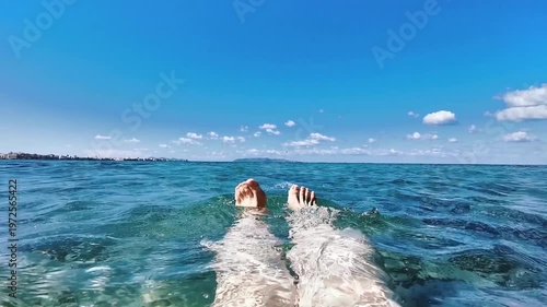 Bare feet of a person floating in clear blue water, with gentle waves and a sunny sky, showcasing a serene beach atmosphere along the coastline