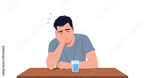 An exhausted man with dark hair leans his head on his hand in a state of fatigue while sitting behind a plain wooden table with a glass of water on a white background.