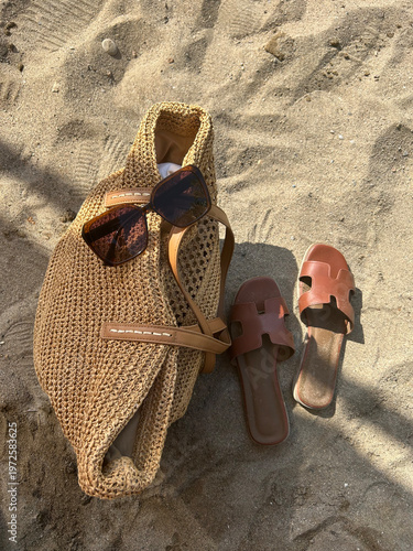 A stylish woven straw tote bag with a eyeglasses, paired with brown leather sandals on sandy beach ground, evoking summer vibes.