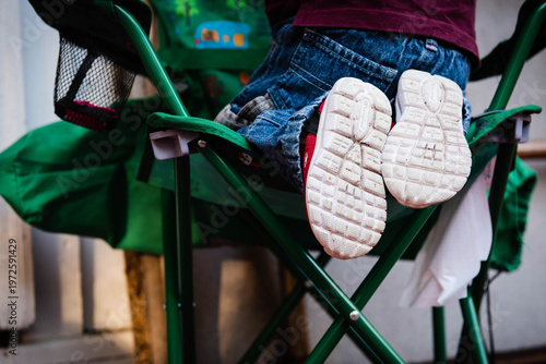 Canvas Print Close-up of a young child's legs in blue jeans and white canvas sneakers dangling from a green folding camping chair, a classic image of outdoor summer childhood