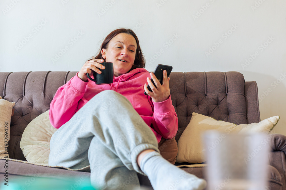 Obraz premium Woman sitting on couch with phone and drink at home in casual setting during daytime