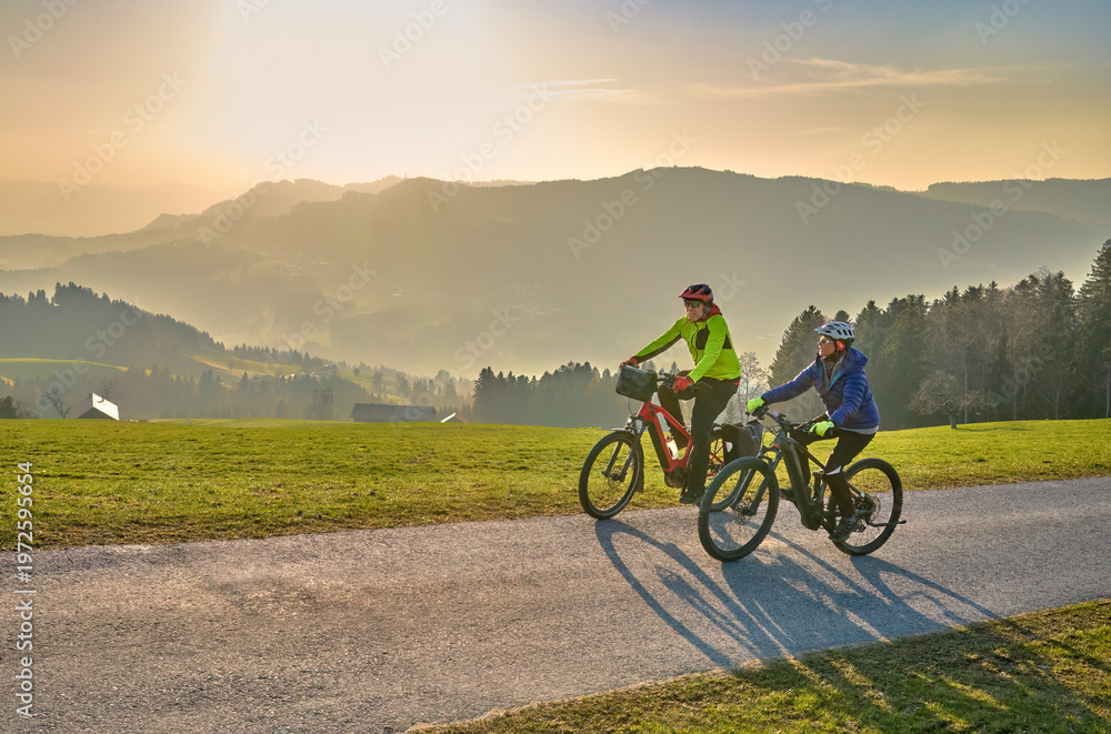 Fototapeta premium Active senior couple riding electric bikes through scenic Bregenzerwald in Austria during golden evening light. Enjoying nature, mountains, and healthy outdoor lifestyle in spring countryside. 