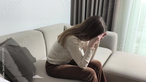 Woman sits on sofa leaning head on hands looking down. Female bends forward resting elbows on knees in sad posture. Scene conveys emotional distress depression and inner mental struggle..