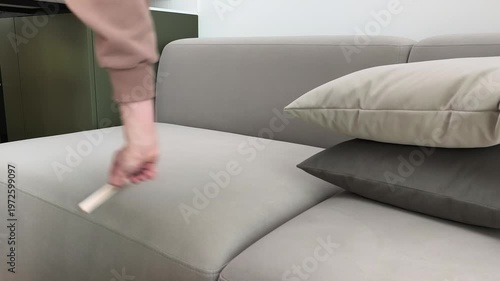 Person cleans velour sofa surface removing hair and dust at home. Hand brushes upholstery carefully maintaining fabric condition indoors