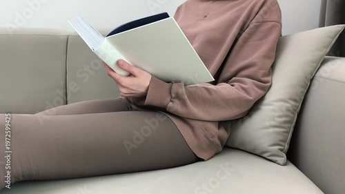Woman flips book pages while relaxing on sofa at home. Female browses content calmly during leisure indoor time. Reading habit and cozy lifestyle concept in apartment living room..