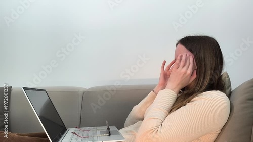 Laptop use continues as woman on sofa covers face feeling sick and low energy. Female without glasses experiences discomfort and apathy during remote work at home. Health problem concept