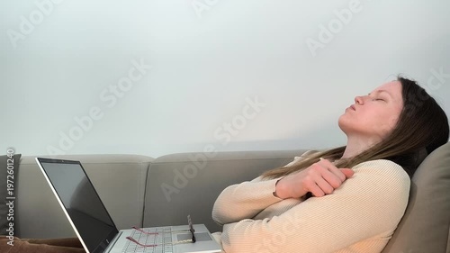 Laptop session stops as woman on sofa leans back closing eyes in exhaustion. Female without glasses breathes out feeling burnout after remote work. Lifestyle concept of fatigue and low