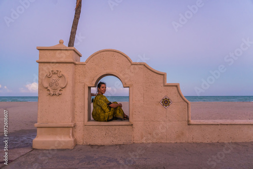 A woman sits in a window of a building overlooking the ocean