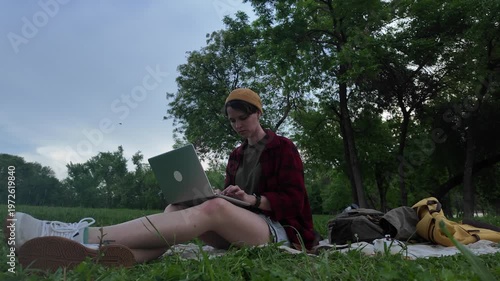 Young woman using laptop and drinking tea while sitting on blanket in green park during evening time. Remote work combined with calm outdoor lifestyle
