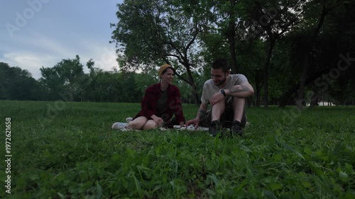 Young man and woman eating chocolate during tea picnic while sitting on blanket in green park in the evening. Couple enjoying calm outdoor moment with snacks and tea
