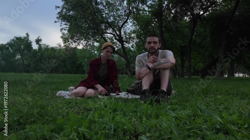 Young man and woman eating chocolate during tea picnic while sitting on blanket in green park in the evening. Couple enjoying calm outdoor moment with snacks and tea