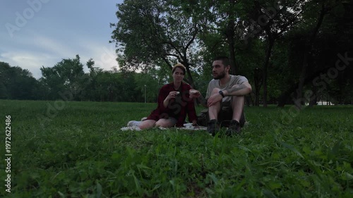 Young man and woman drinking Chinese tea from small cups while sitting on blanket in green park in the evening. Couple enjoying calm conversation during outdoor tea picnic